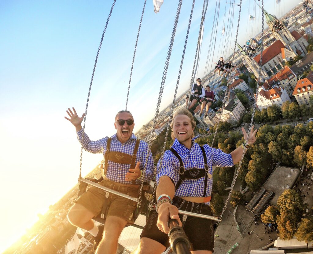 two young men wearing lederhosen and blue and white checkered shirts ride The Wellenflug at Oktoberfest and snap a selfie with a Selfie Stick.