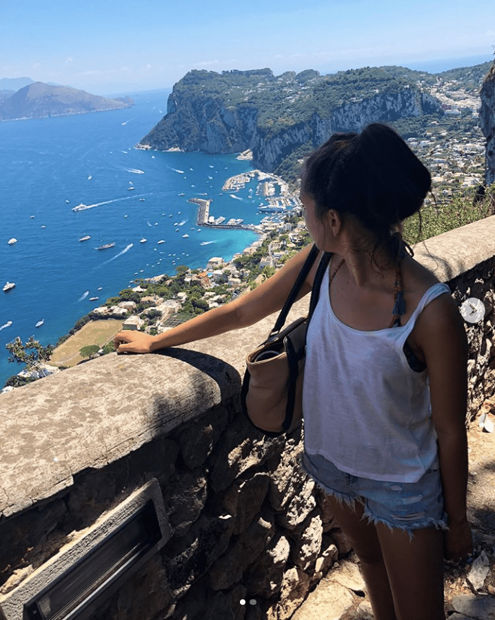 a young woman peers over the view from the Phoenician Steps. She wears jean shorts and a loose white tank top. The water is bright blue and boats can be seen coming in and out of Marina Grande on a sunny day.