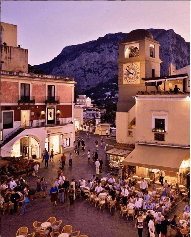 image of La Piazzetta in Capri. The piazza is full of dining locals and tourists, sitting in wicker chairs around small tables. It is dusk and the sky is pink. The lights around the piazza illuminate the clock and the buildings